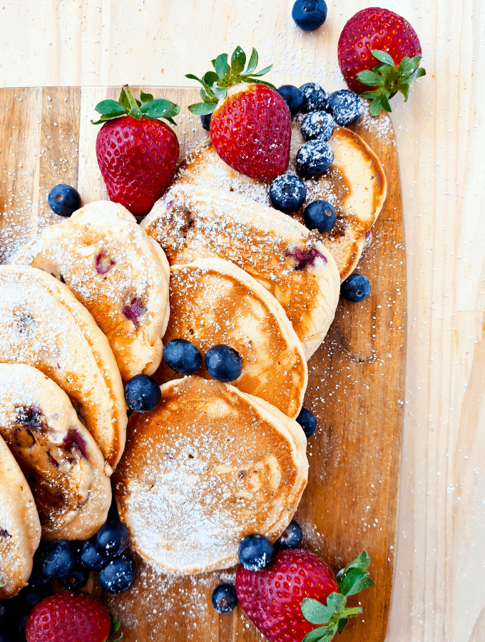 Pancakes sprinked with icing sugar/powder on top of a wooden board, decorated with blueberries and strawberries