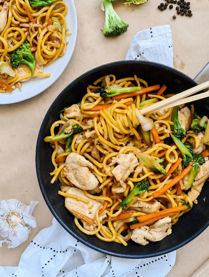 Freshly cooked chicken with noodles stir fry and vegetables in a non-stick pan. Fresh garlic and a tea towel in the background