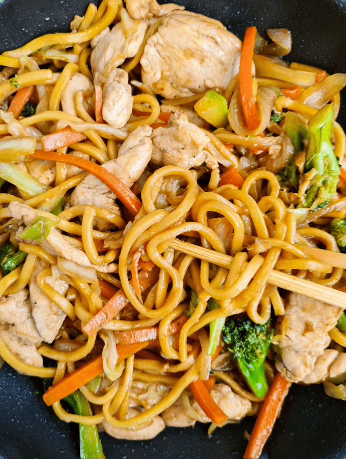 Close up of noodles, chicken breasts and broccoli in a frying pan. A pair of chopsticks picking up noodles
