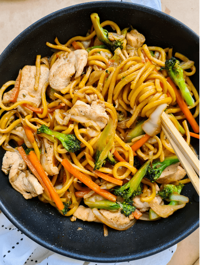 A pair of chopsticks twirling up hokkien noodles in a pan full of cooked noodles, chicken and vegetables