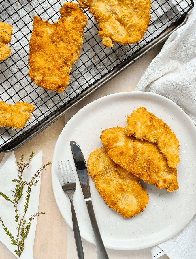 Golden crispy panko coated chicken schnitzel pieces on a plate and some on a cooling rack.