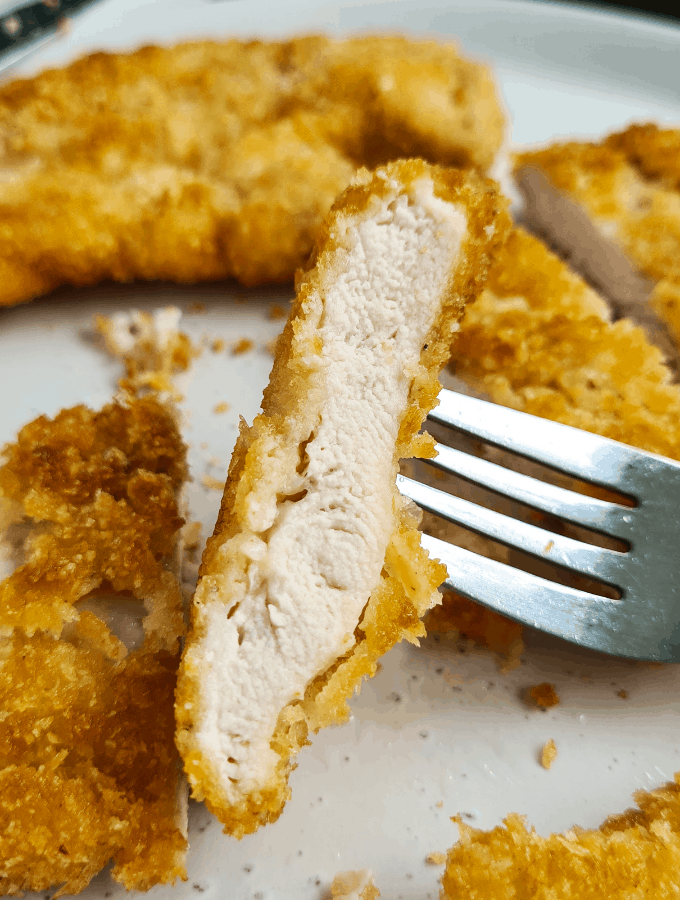 close up view of a sliced piece of panko chicken schnitzel on a fork. Surrounded by pieces of panko chicken schnitzel