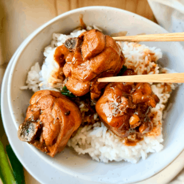 3 pieces of soy sauce chicken sat on top of a bowl of rice with sauce drizzled on rice. chopsticks holding onto 1 piece of chicken