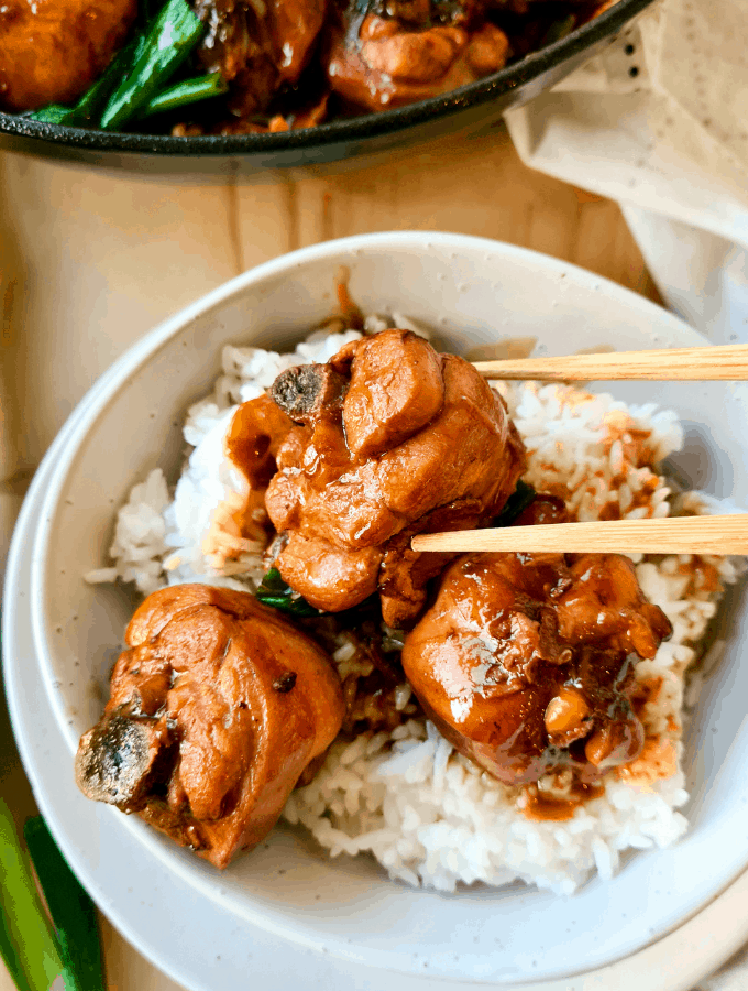 Bowl of rice with 3 pieces of soy sauce chicken sat on top. Sauce drizzled on rice. Chopsticks holding onto 1 piece of chicken.