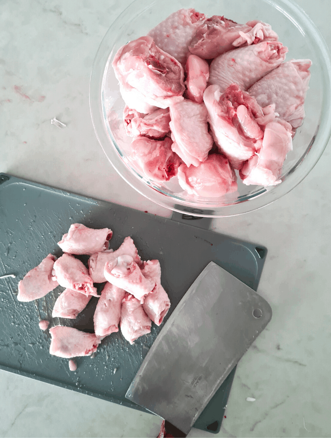 chopping board with chicken knee caps chopped off drumsticks. A thick bladed cleaver resting on chopping board. A bowl of chopped chicken drumsticks