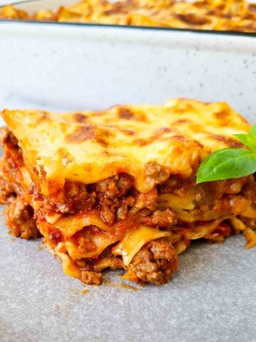 Sideview of a slice of lasagna showing all 4 layers. A tray of lasagna in the background, blurred