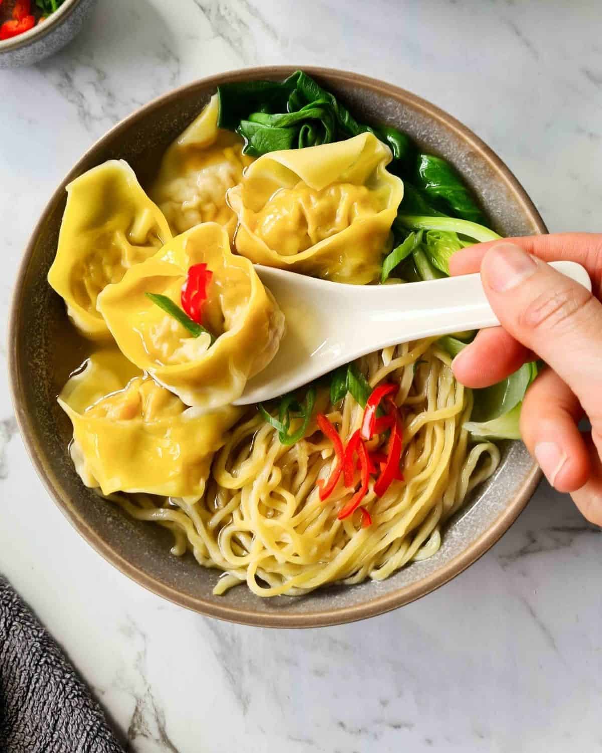 close up of a wonton with soup in a soup spoon being scooped out of a bowl