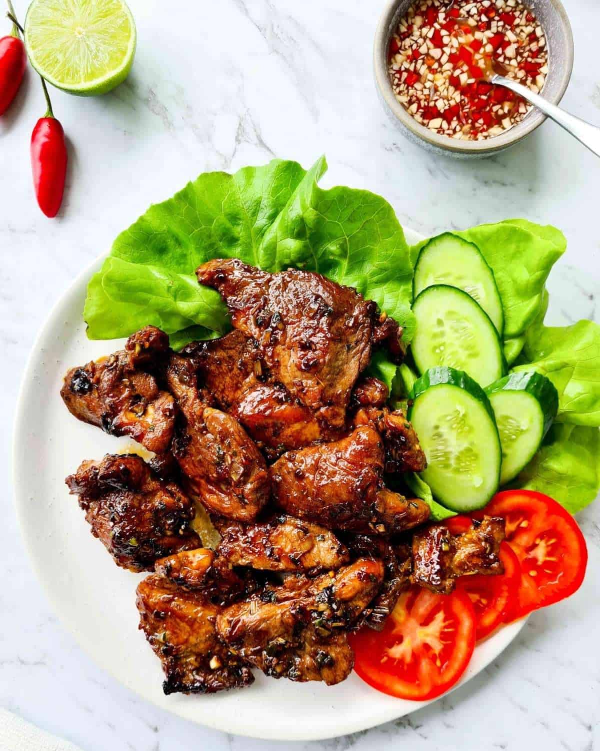 A large plate of cooked Vietnamese pork chops with fresh lettuce, cucumber and tomato slices.