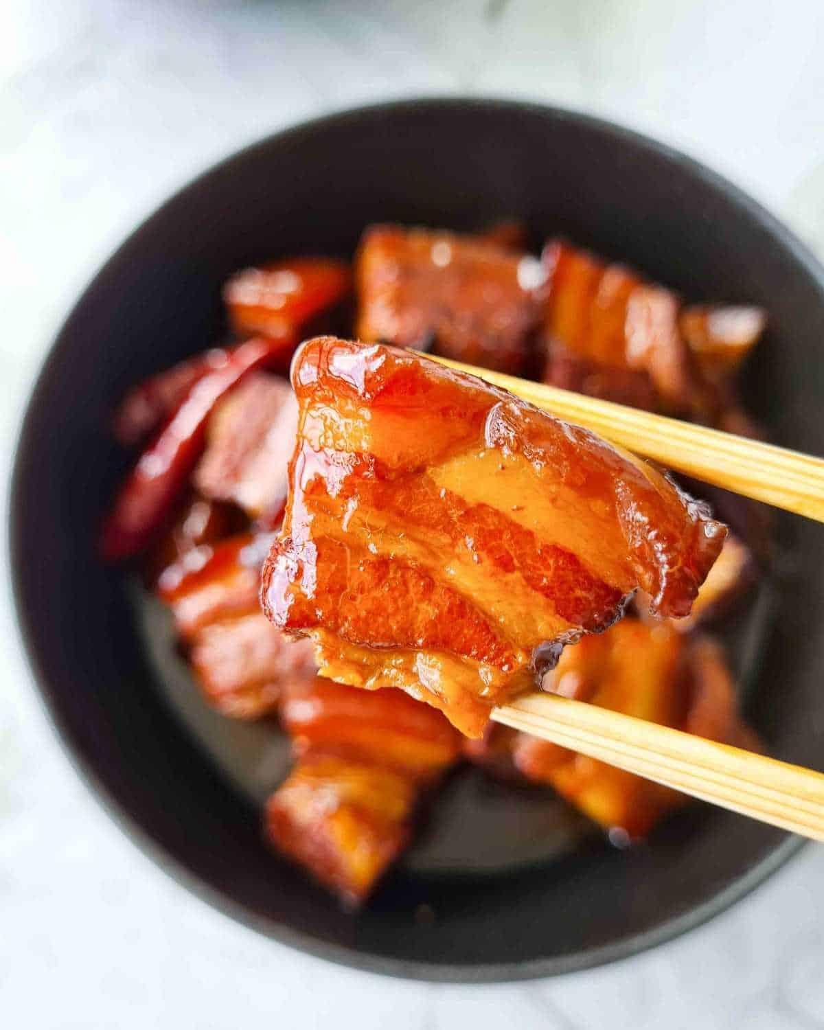 Close up of a piece of glossy pork belly being held up by a pair of chopsticks