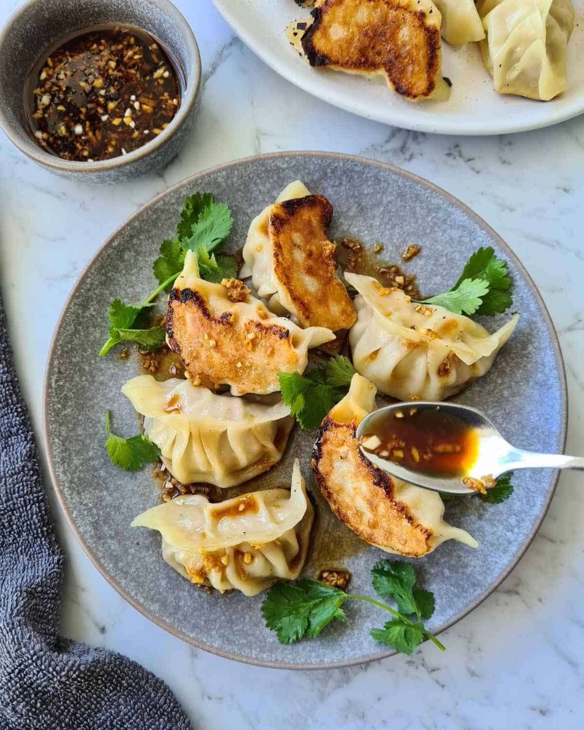 Cooked dumplings on a small grey plate with dipping sauce being drizzled over with a spoon