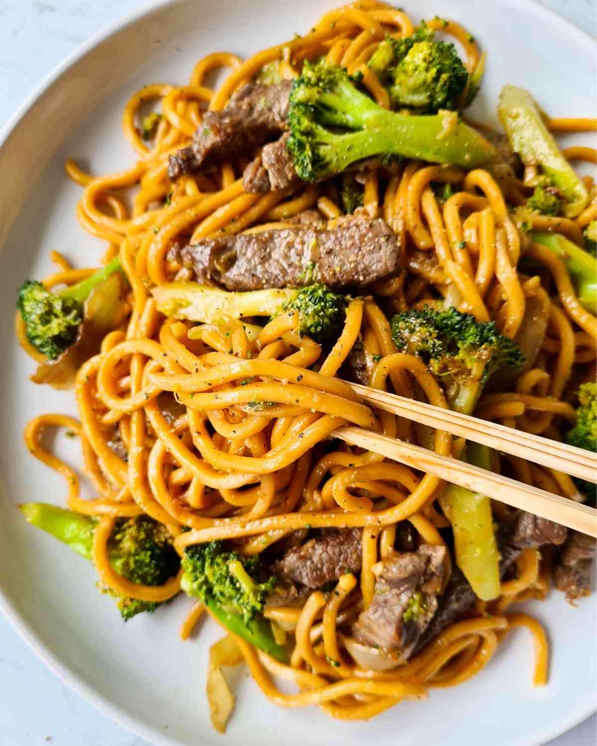 Stir fried noodles with broccoli, beef strips on a large white plate and a pair of chopsticks