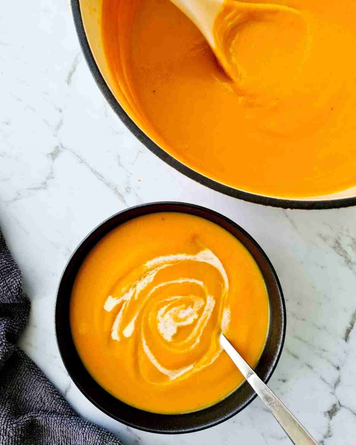 A bowl of smooth pumpkin and sweet potato soup with the pot of soup in the top right hand corner